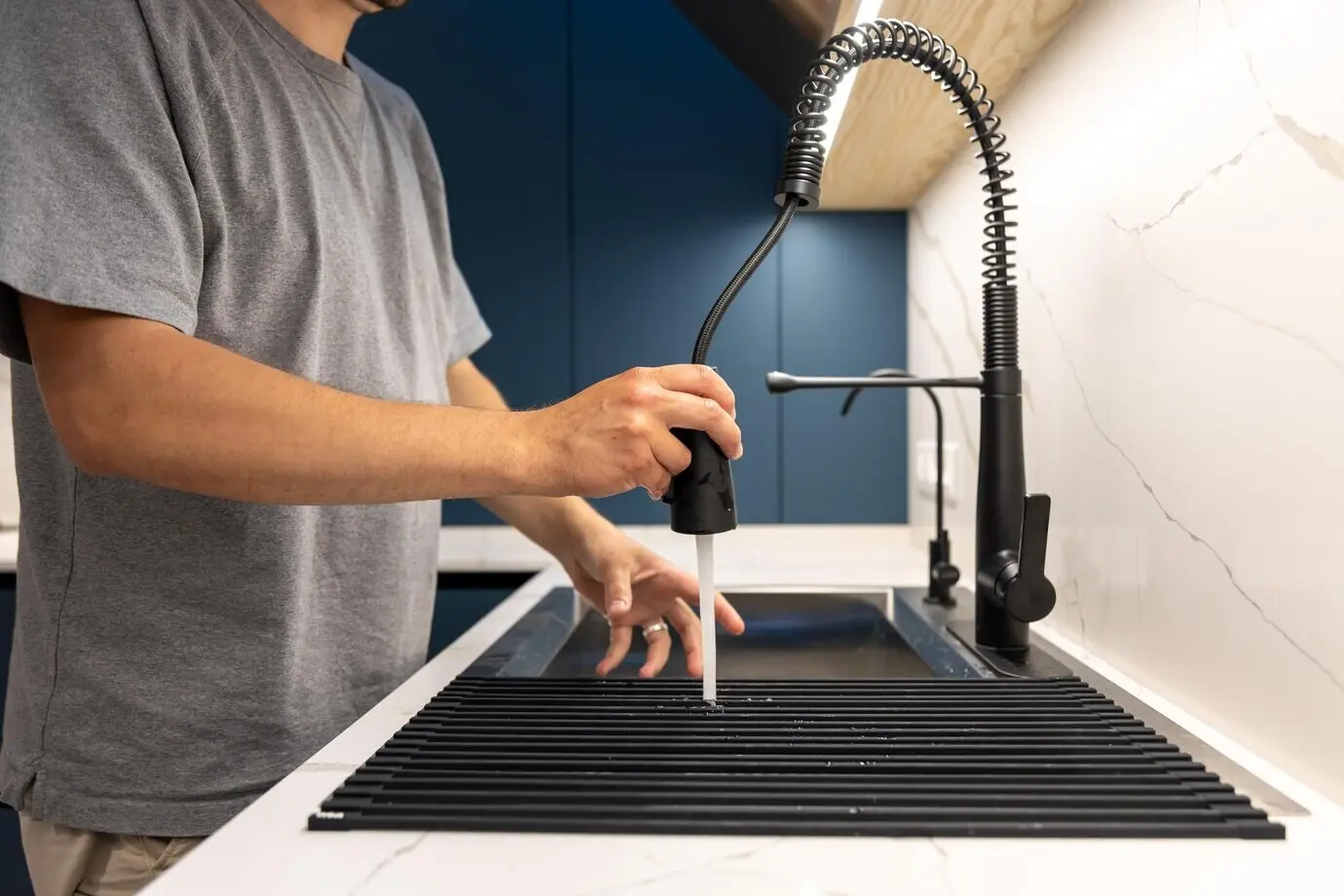 A man uses a pull-out faucet sprayer to clean a kitchen rack, rinsing the drainer.
